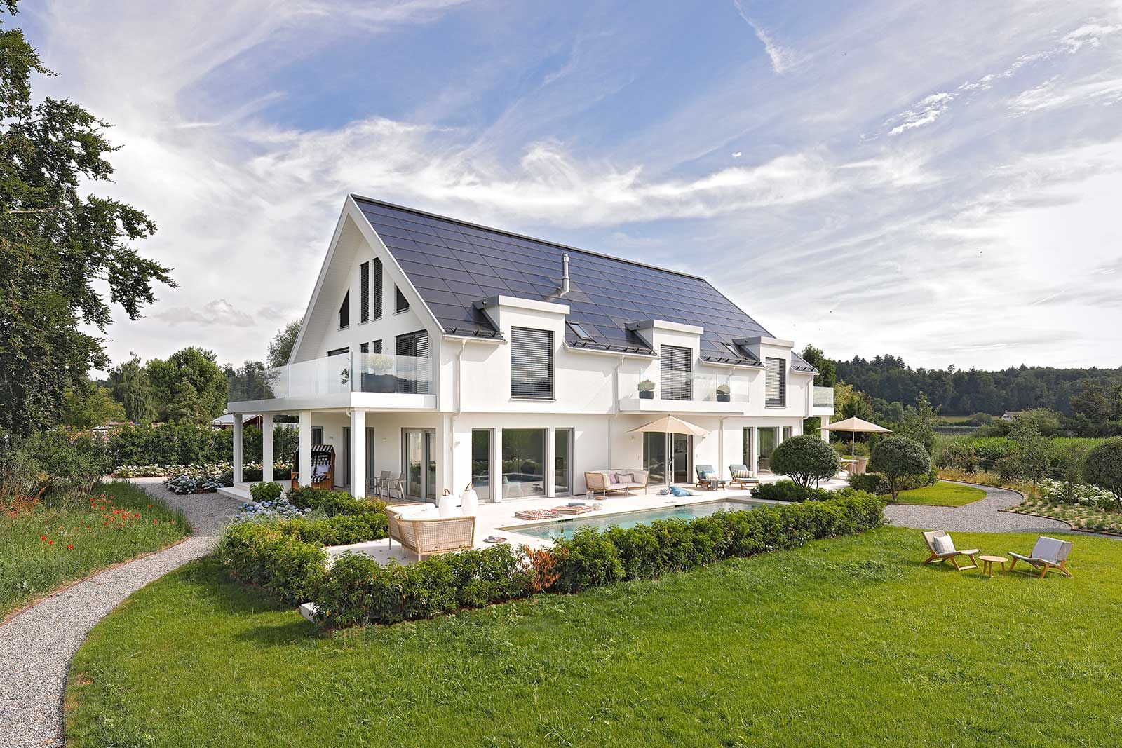 Glatte Dachziegel und eine flächige PV-Anlage verkörpern moderne Klarheit und Nachhaltigkeit. Sie treffen auf traditionelle Anleihen wie dem Satteldach und die seitlichen Gauben. Die weissen Putzfassaden werden durch grosszügige Glasflächen ergänzt.