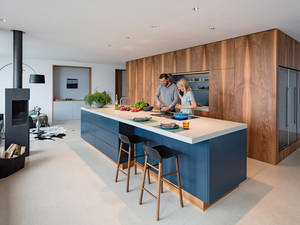 Hinter den echtholzfurnierten Fronten verstecken sich moderne Geräte und ein begehbares Reduit. Davor ist die dunkle Inselküche ein echter Blickfang. Herzog Küchen.