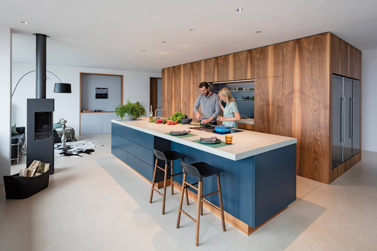 Hinter den echtholzfurnierten Fronten verstecken sich moderne Geräte und ein begehbares Reduit. Davor ist die dunkle Inselküche ein echter Blickfang. Herzog Küchen.