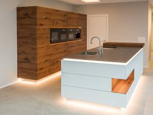 Durch die Sockelbeleuchtung und den verspiegelten Sockel wirkt die Küche, die durch die Fronten aus Altholz-Eiche besticht, als würde sie schweben. Alpnach Küchen AG.