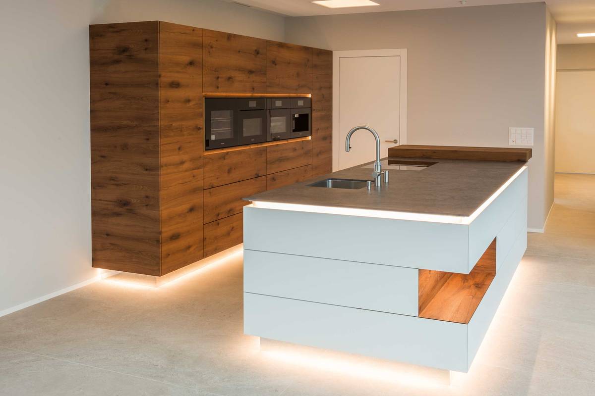 Durch die Sockelbeleuchtung und den verspiegelten Sockel wirkt die Küche, die durch die Fronten aus Altholz-Eiche besticht, als würde sie schweben. Alpnach Küchen AG.
