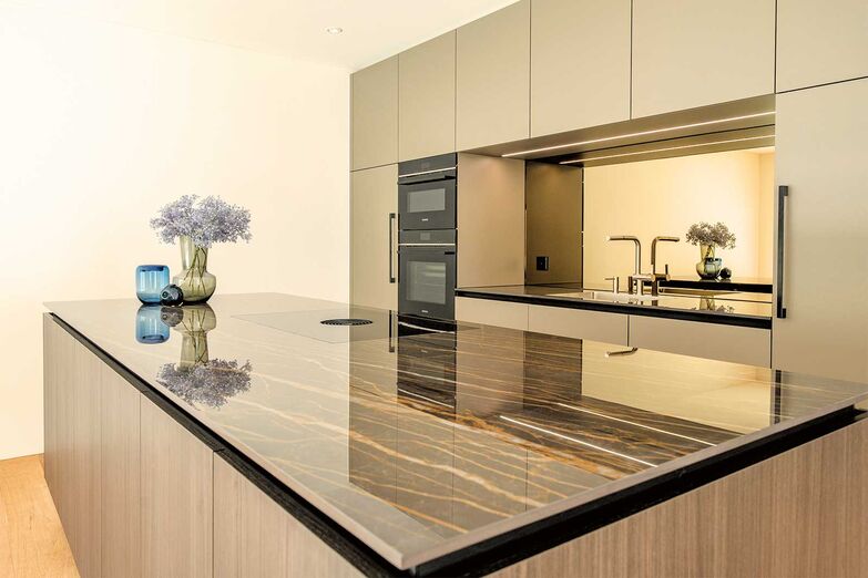 Tiefe und Helligkeit: Diese stilvolle Küche vereint modernes Design mit funktionalen Elementen. Die Fronten in Achatgrau der «X-Line»-Griffleiste wirken elegant und zeitlos. Ein Highlight ist die polierte Keramik­abdeckung «Noir Desir», die mit ihrem dezenten Glanz edle Akzente setzt. Eindrucksvoll ist die bronzefarbene Spiegelrückwand, die Licht reflektiert und dem Raum Helligkeit und Tiefe verleiht.   Hans Eisenring AG, www.eisenring-kuechenbau.ch