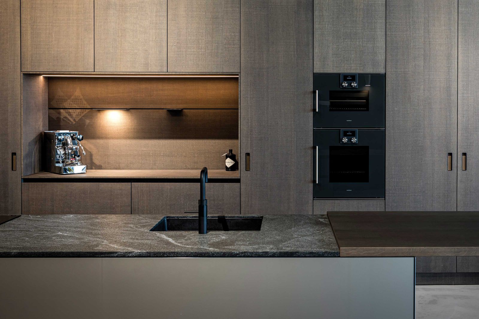 «Orea +» mit sägerohen Eichenfronten und einer Insel mit Natursteinabdeckung und Fronten in glänzendem Alucobond.