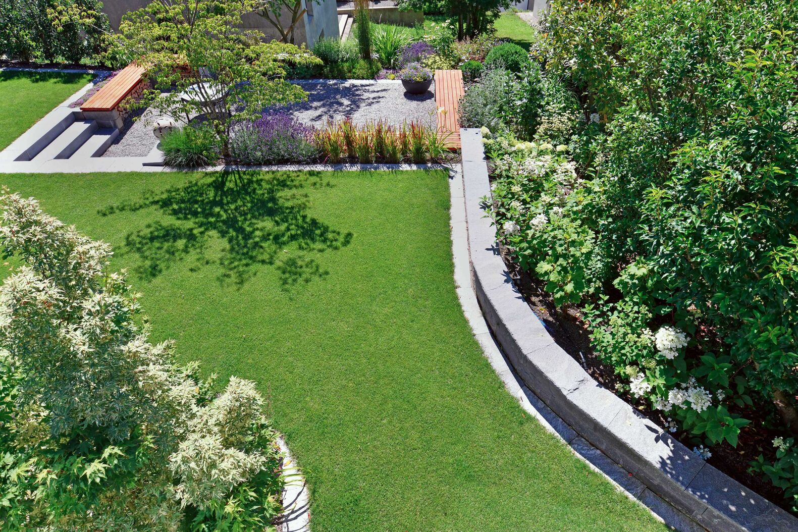 Gartenplanung Jost Gartenbau: Die Mauer aus Maggia-Steinen und eine Hecke aus Burkwoods-Duftblüte rahmen diesen Garten ein, der mit vielen Elementen und Details neu gestaltet wurde und rundum begeistert. Im Hintergrund lädt ein grosszügiger Grillplatz zu gemütlichen Stunden um die Feuerschale ein, bei den Pflanzen setzen Blutgras und Purpurglöckchen rote Akzente, Rispenhortensien sorgen das Jahr hindurch für wechselnde Farbtupfer. jostgartenbau.ch