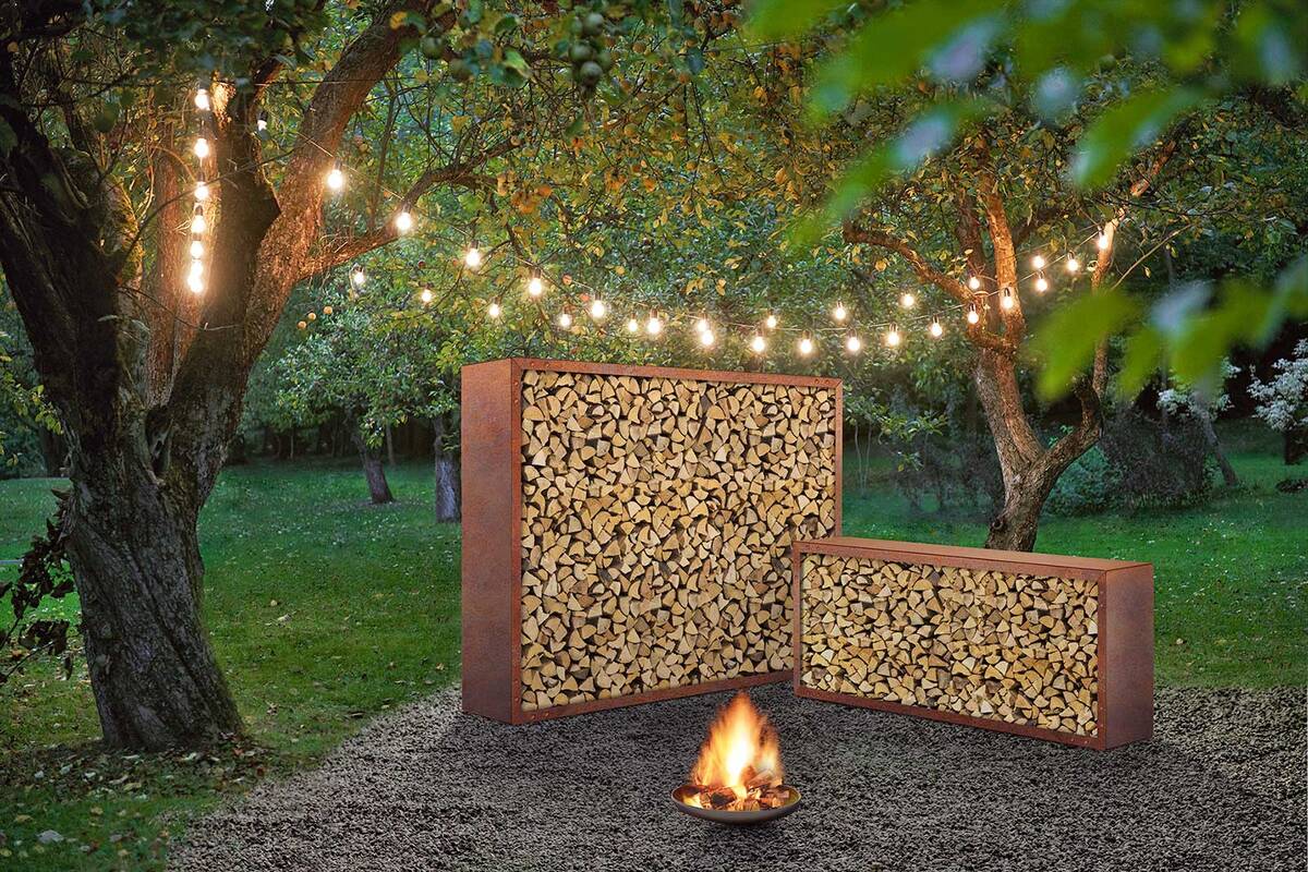 Eine Feuerschale inmitten eines Waldstücks, dazu ein Holzlager, beleuchtet und in Szene gesetzt. Sager steht für hochwertige Gartenobjekte, die individuell auf Mass angefertigt werden oder auf Standardgrössen basieren. Sager Gartengalerie AG.