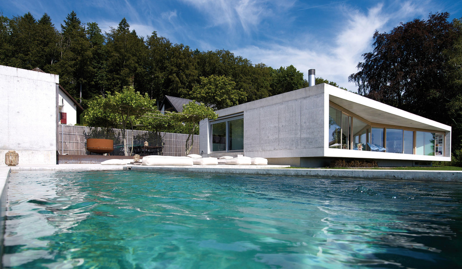 Sichtbetonschwimmbad an Hanglage, mit chlorfreier, geruchsneutraler Wasseraufbereitung mit Aktivsauerstoff, automatischer Unterflur-Rollladenabdeckung und PVC-Solarlamellen mit Rollladenparkkasten. AC Schwimmbadtechnik AG.