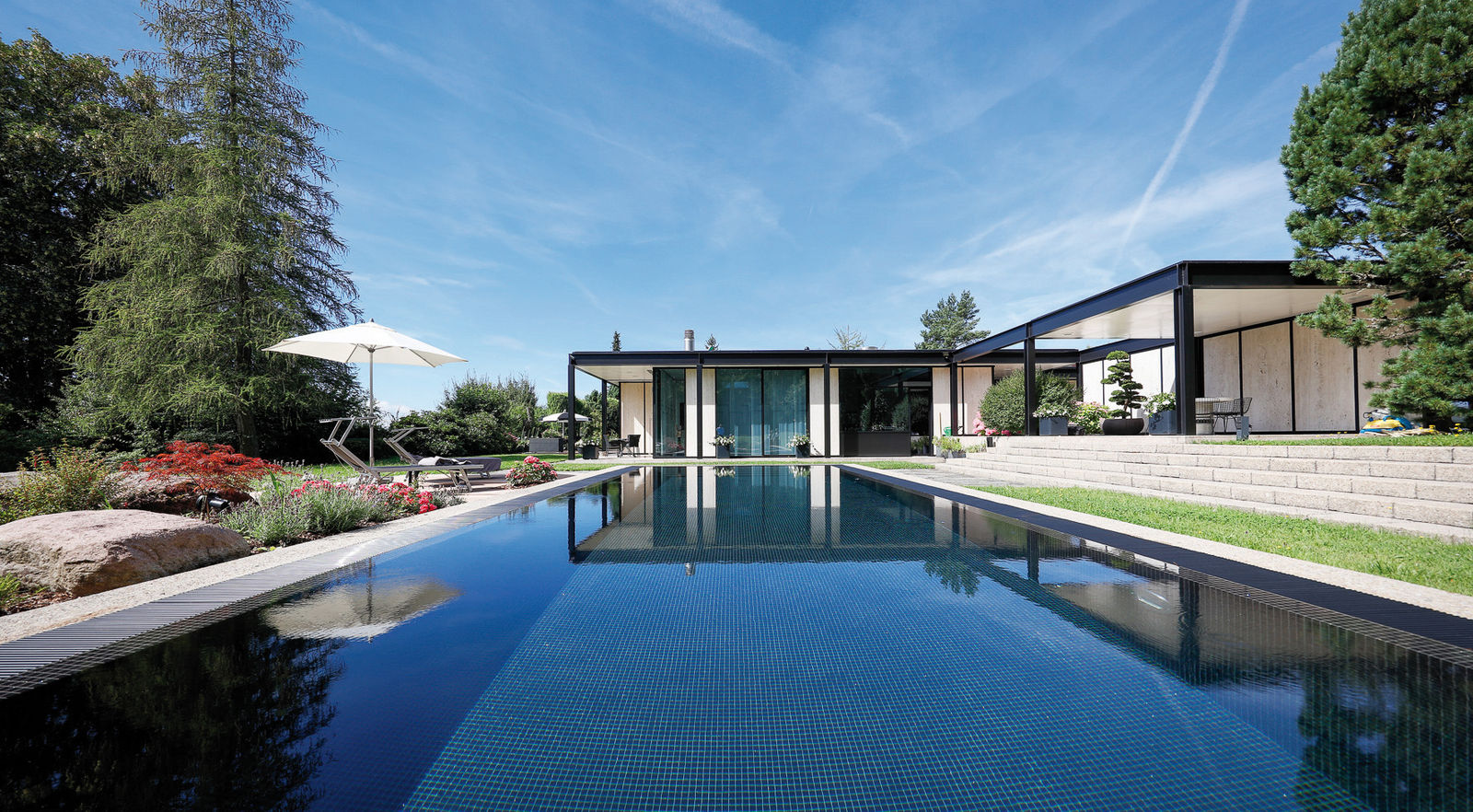 Der schwarze Überlaufpool wurde in die bestehende Beckenumrandung aus Waschbeton integriert. Dazu passend der Abdeckrost mit seiner hohen Schwallwasser-Aufnahme sowie das wetterfeste Holzdeck, das sich als Ruheoase anbietet und unter dem die komplette Technik untergebracht ist. 
