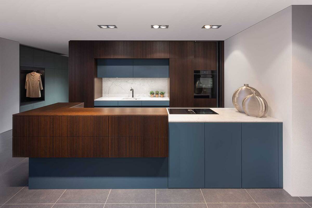 Elbau kombiniert dunkle Räuchereiche und petrolblaue Fronten. Aufgebrochen werden die dunkleren Elemente mit einer Abdeckung und einem Becken aus italienischem Naturstein. Dieser wird mit einer LED-Beleuchtung noch stärker hervorgehoben und betont die natürliche Marmorierung des Steins. Elbau Küchen AG.