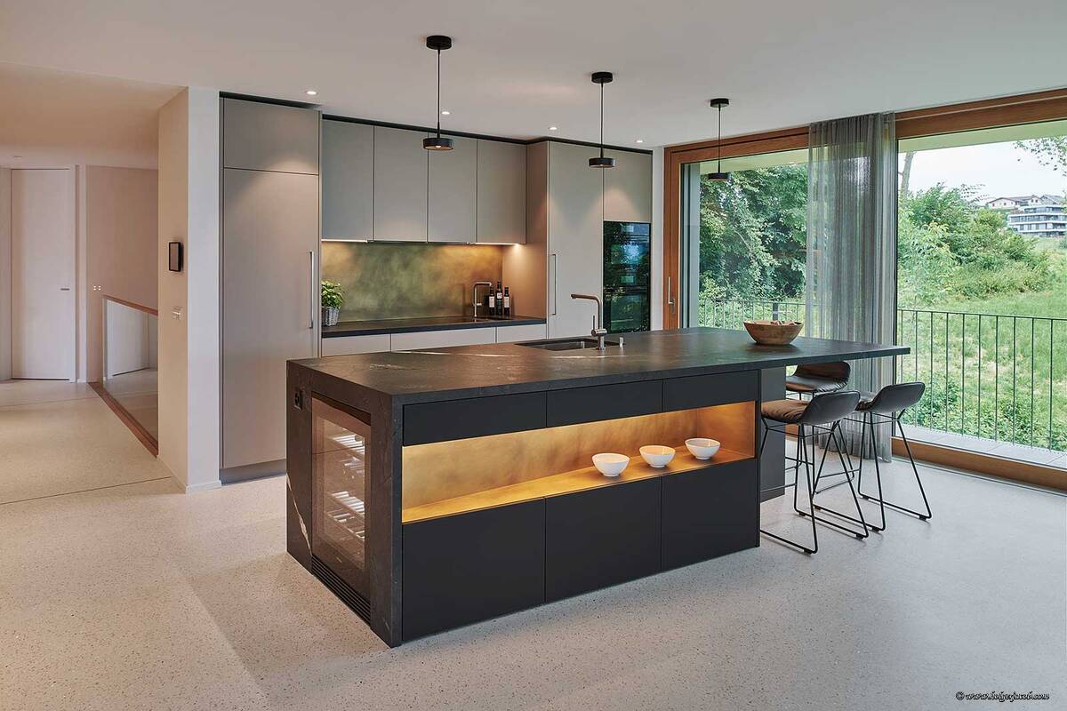 Ein Bijou mit Ecken und Kanten: Die schwarze Kochinsel harmoniert mit den Hochschränken im eleganten Naturgrau. Messing-Rückwände und die lederne Oberflächenstruktur des Negresco-Granits runden die edle Küche ab. Brunner Küchen.