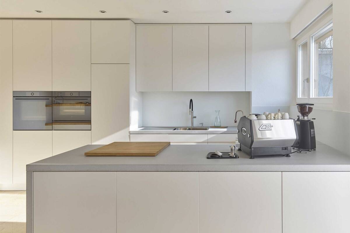 Die helle Küche besticht durch ihre Abdeckungen und Seiten aus Echtbeton. Die Fronten sind spritzlackiert. Das Wandboard schenkt zusätzlichen Platz für Küchenutensilien. Oesch Innenausbau AG.
