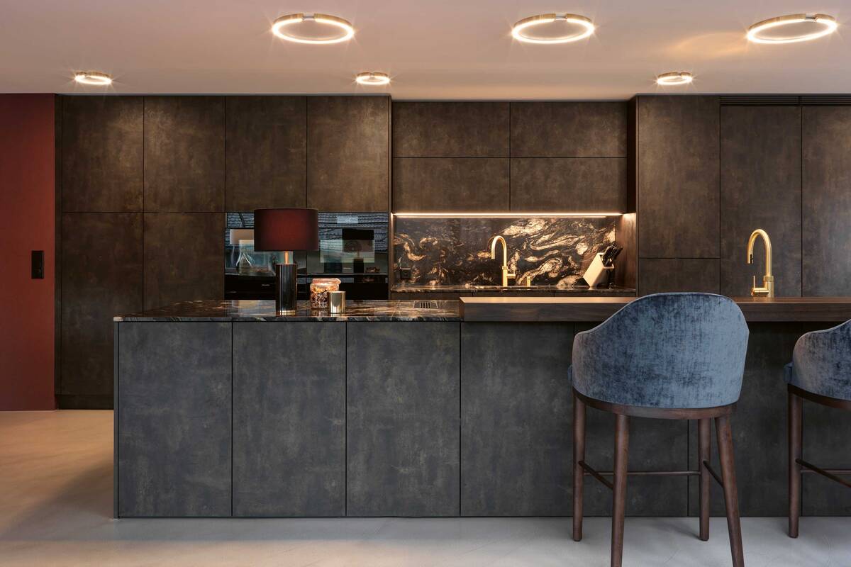 Die Küche kombiniert Kunstharzfronten mit einer Granitabdeckung und einer Theke in Räuchereiche. Integrierte LED-Spots sowie Armaturen in Messing-Optik schaffen ein warmes Ambiente. Brunner Küchen AG.